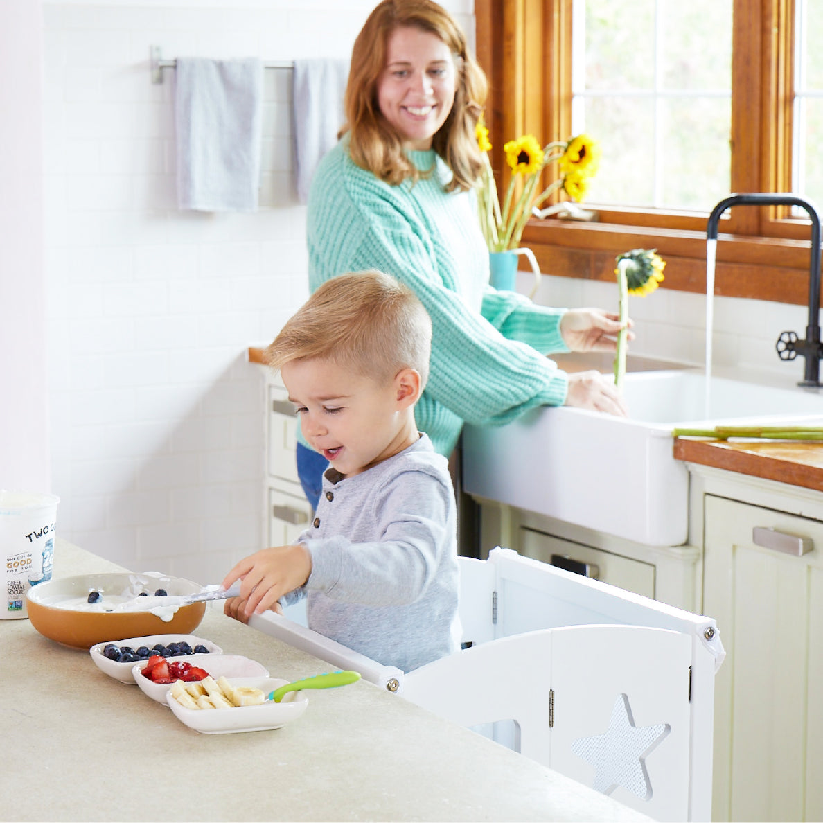 Image of toddler boy in the a Classic Kitchen Helper - White making breakfast with his mother in their kitchen. 