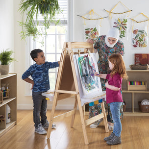 A teacher watches as a boy and girl paint on an easel in a bright classroom with nature decorations.