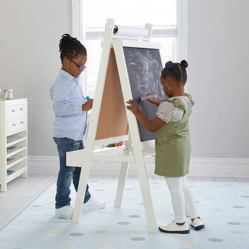 Two young children draw together on a chalkboard easel in a bright, modern playroom.