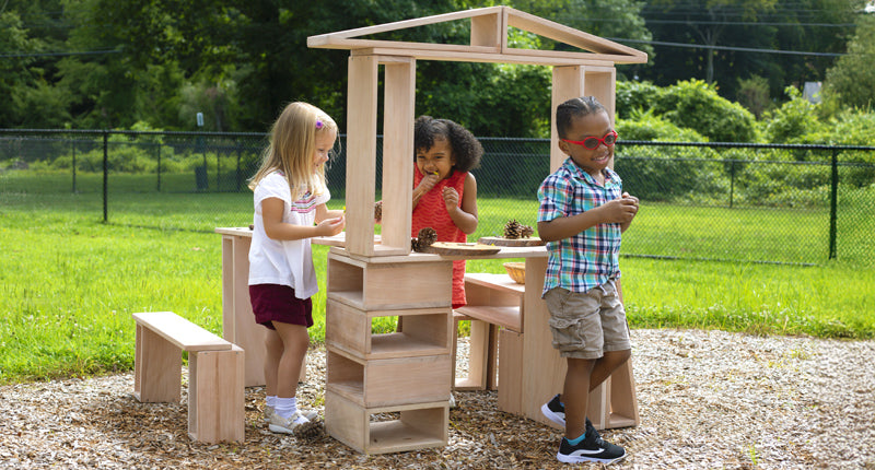 Three young children play and smile around a wooden playhouse outdoors on a sunny day.