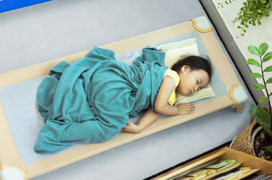 A young child sleeps on a cot with a teal blanket, surrounded by books and green plants.