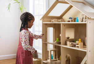 A young girl plays with a wooden dollhouse filled with miniature furniture and figures by a window.