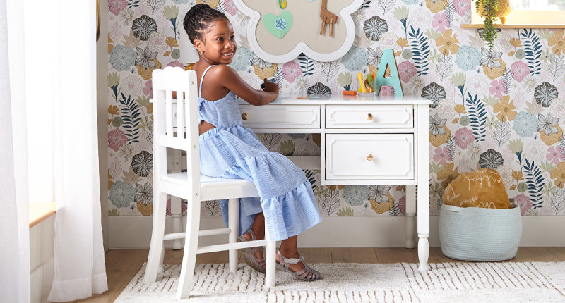 Fashionable young 6 year old girl sitting at a white desk with brushed gold drawer pulls and turned legs in a room with tasteful floral wall paper
