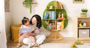 A mother and young child sitting in her lap reading a book, sitting next to a green and natural tree shaped bookshelf