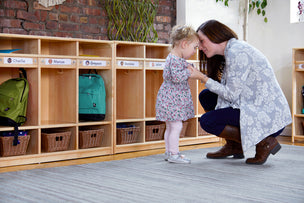Woman kneeling to comfort a young girl in front of cubbies in a classroom or daycare setting.