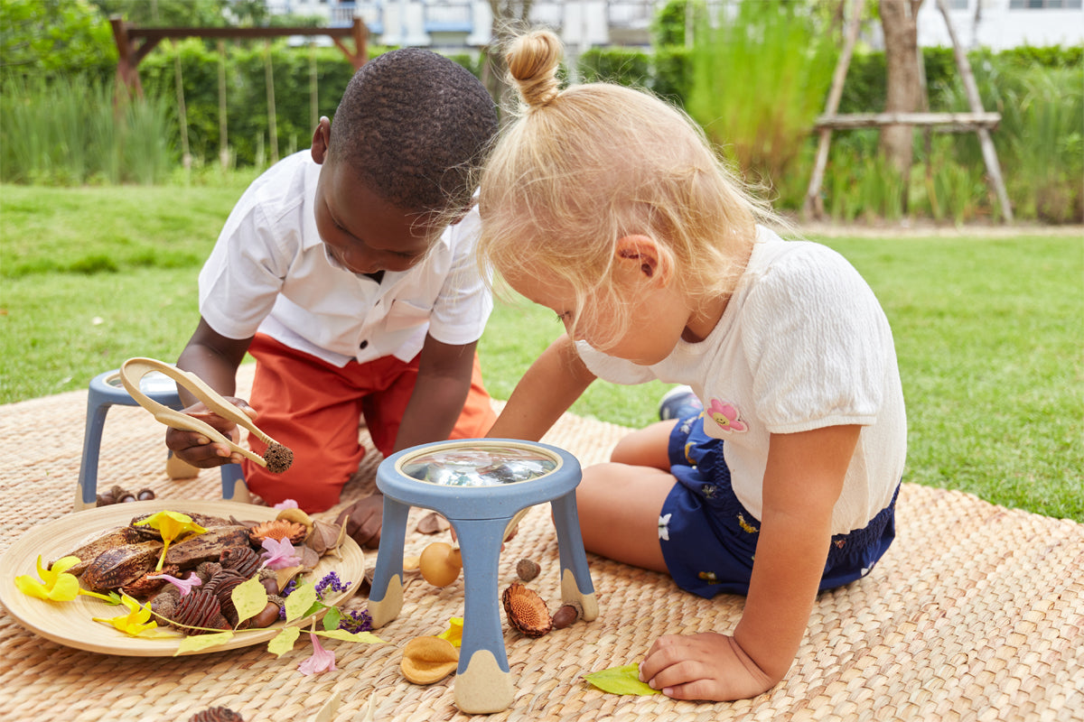 Two young children explore natural objects on a mat outdoors using wooden tongs and magnifying glasses.