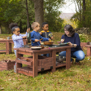 A teacher and three young children play and explore nature at a wooden outdoor activity table in a grassy area.