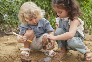 Two young children digging and brushing rocks in sand, possibly searching for fossils, outdoors.