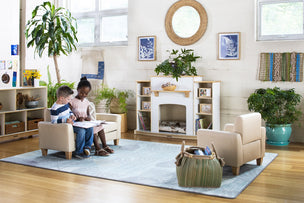 Two children sit together reading on small armchairs in a cozy, light-filled classroom with plants and books.