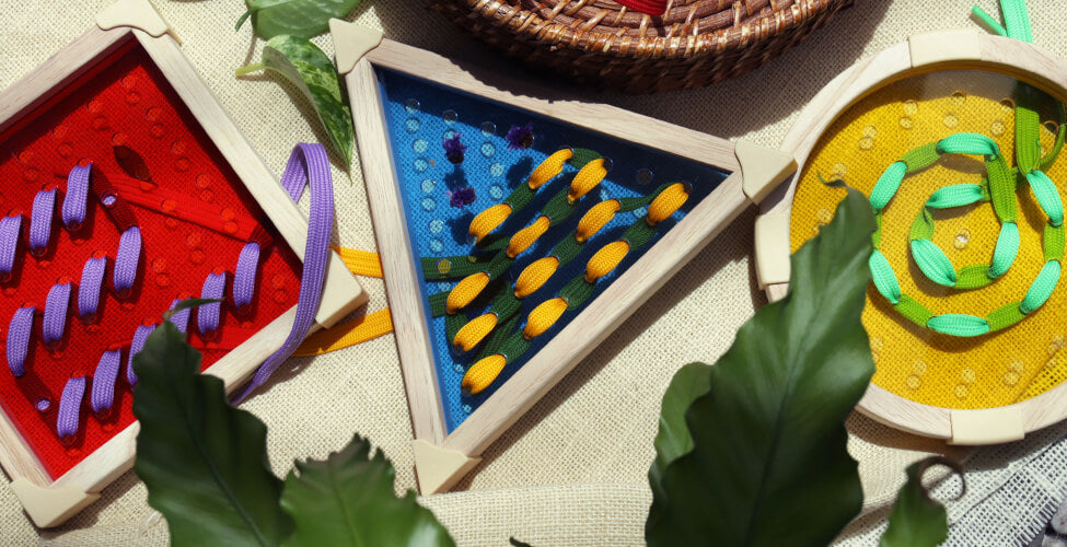 Colorful geometric wooden lacing toys with thread and beads, surrounded by green leaves and a wicker basket.
