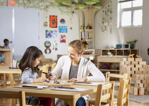 A teacher and a young girl sit at a table in a bright classroom, working on an activity together.