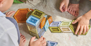 Children playing on the floor with colorful magnetic tiles, building shapes with their hands and feet nearby.