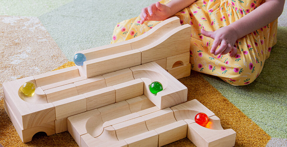 A child in a yellow dress plays with a wooden marble run set on a carpeted floor.