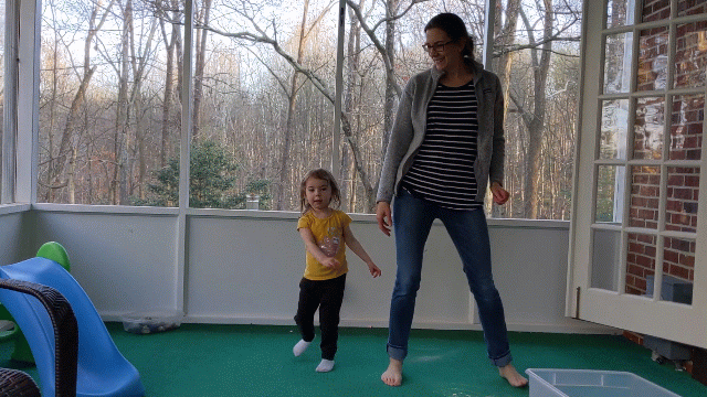 A joyful woman and a young girl dance together on a screened porch, surrounded by the gentle glow of natural light and views of lush trees outside.