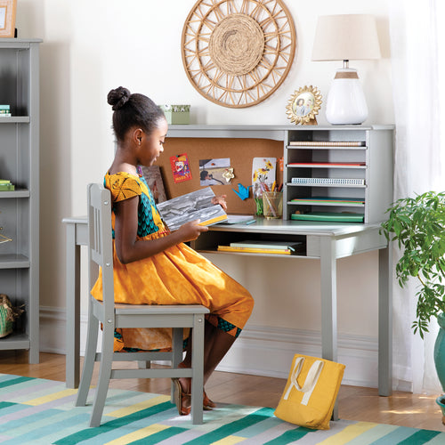 A young girl in a colorful dress reads at a gray desk with shelves, surrounded by books in a bright, tidy room.