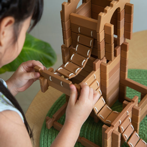 A child builds a wooden toy castle with bridges on a table, assembling the pieces atop a green play mat.