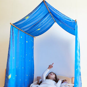 Child lying in bed under a blue Double Playsilk canopy decorated with stars and planets, pointing upward.