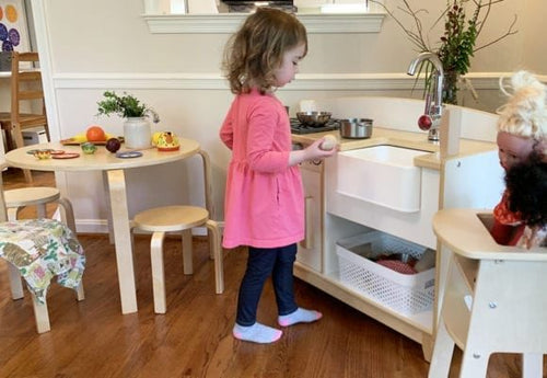 A young girl plays at a toy kitchen set in a room with wooden floors and small tables, pretending to cook delightful meals.