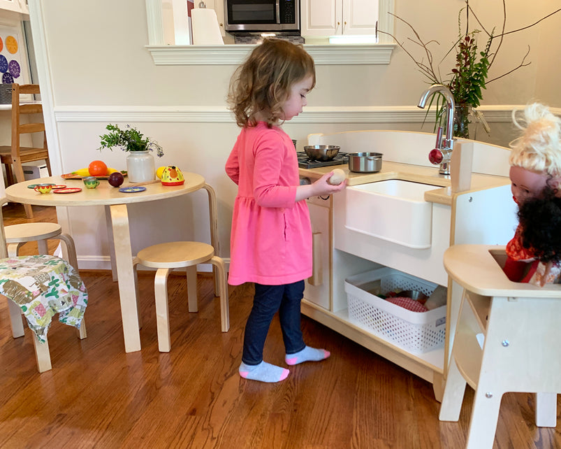 Young girl playing in a toy kitchen, with play food and a doll nearby, on a wooden floor, creating an imaginative scene.