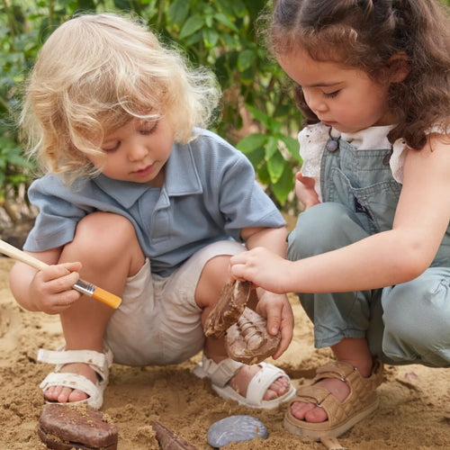Two young children use brushes to carefully examine toy dinosaur fossils as they dig in the sand outdoors.