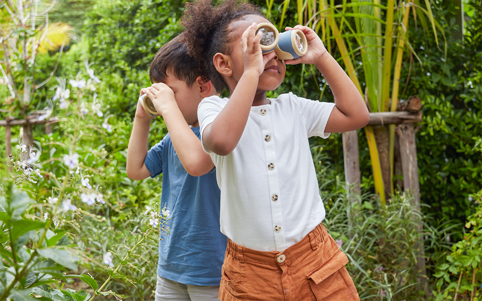 Two children in a garden explore nature using homemade binoculars crafted from cardboard tubes.