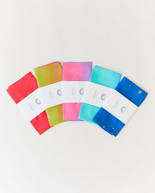 Five colorful folded Playsilk cloths arranged in a fan shape on a white background, each with a branded label. Made from natural materials, these open-ended toys inspire creative play and imagination.