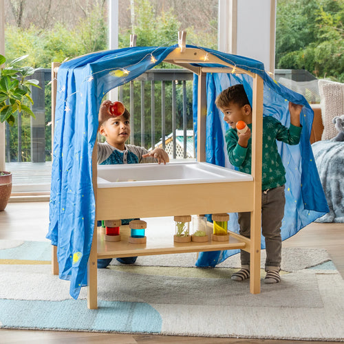 Two young children play under a blue fabric canopy over a wooden sensory table, exploring textures and colors indoors.