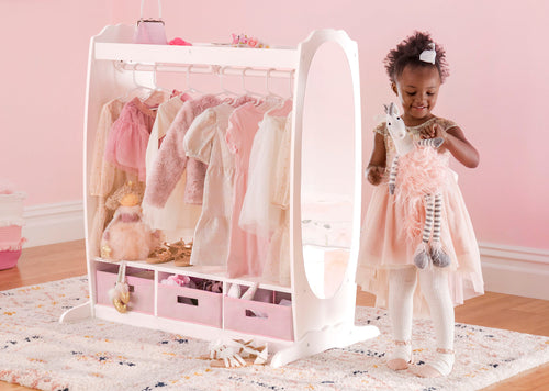 A young girl in a pink dress hugs her stuffed toy beside a colorful kids’ wardrobe filled with dresses and toys.