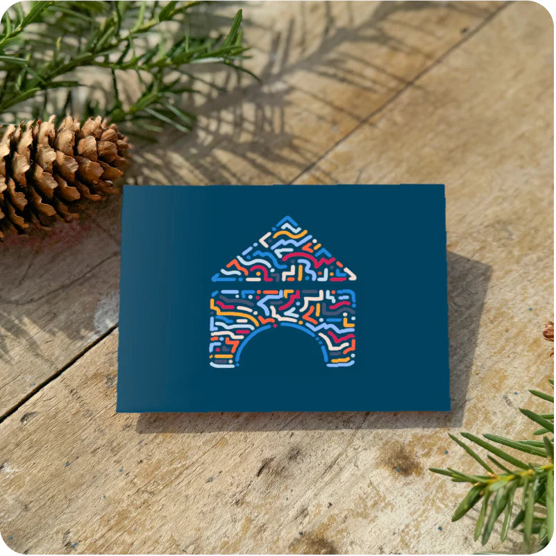 A festive card with a colorful house design rests on a wooden surface, surrounded by pine branches and a pinecone for a cozy holiday touch.