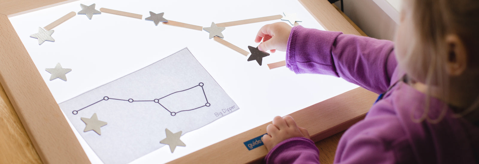 A child arranges star shapes on a light table, forming the Big Dipper constellation and exploring patterns in a creative, hands-on way.
