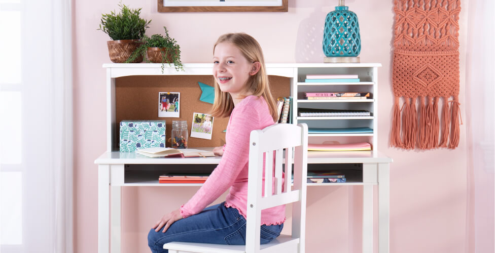 A smiling girl in a pink shirt sits at a white desk with books and plants, surrounded by soft pink decor.