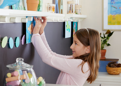 A girl arranges vibrant artwork on a wall shelf in a cheerful, art-filled room.