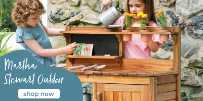 Two children playing with a wooden outdoor play kitchen set against a stone wall.