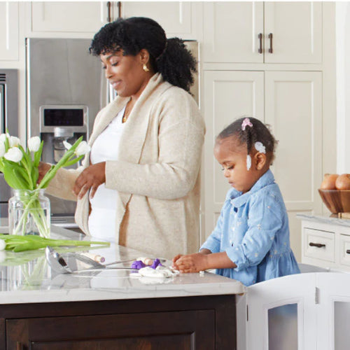 A woman arranges fresh flowers while a young girl plays at a sunny kitchen counter.