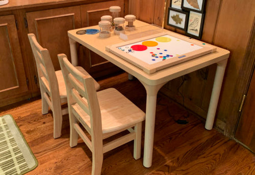 Two wooden chairs and a table with paint jars and art supplies on a hardwood floor beside spacious wooden cabinets.