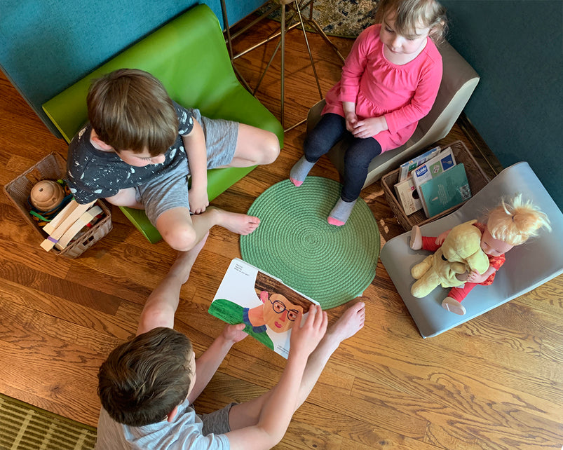 Three children sit on chairs around a green mat, exploring a colorful picture book together on a wooden floor.
