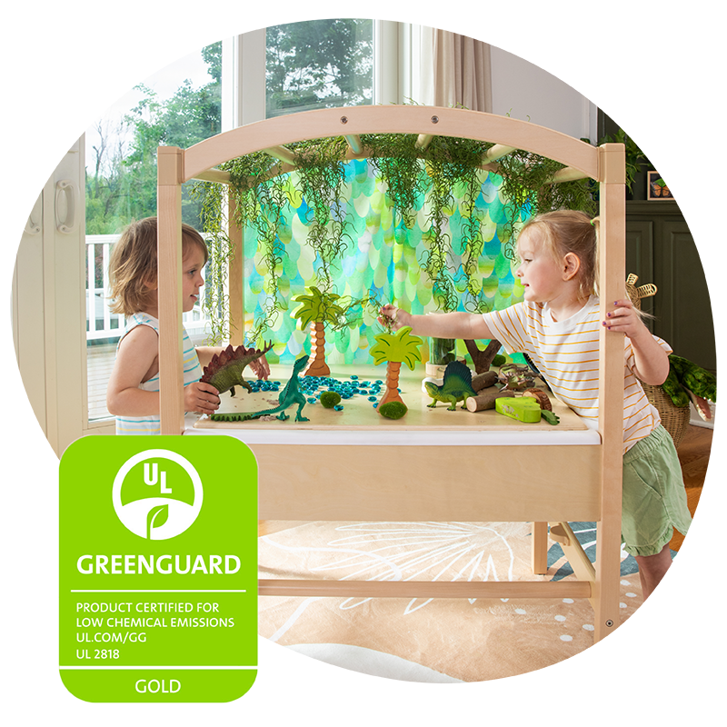 Two young children explore their imagination with dinosaur toys at a wooden activity table surrounded by trees and plants.