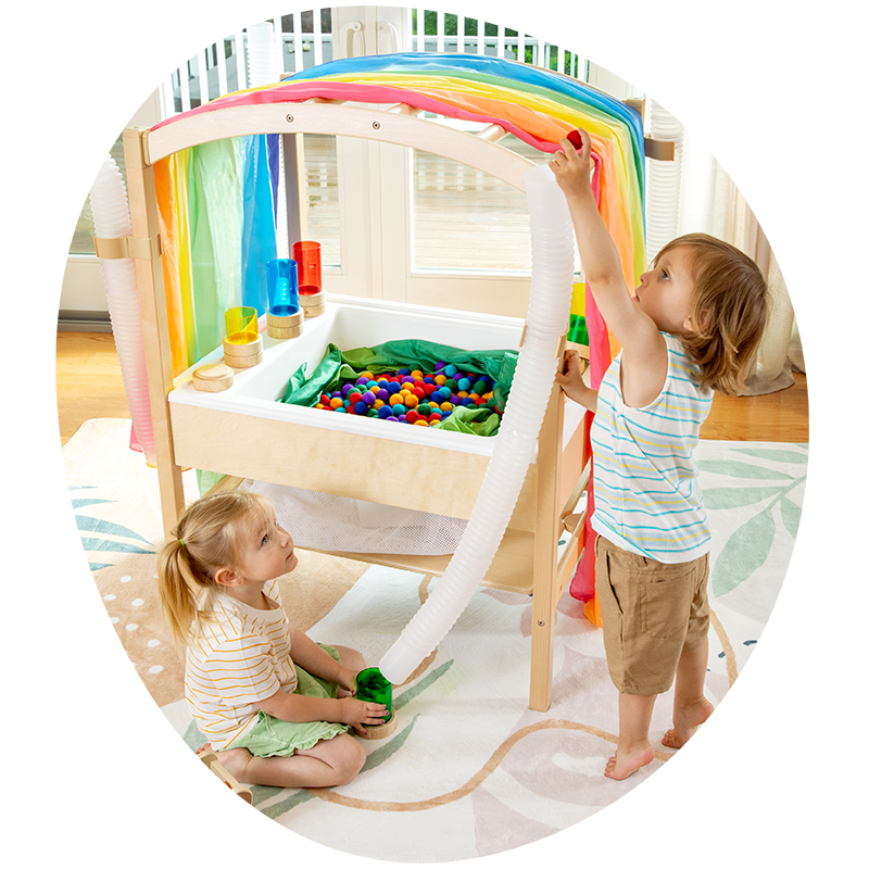 Two young children explore a sensory table draped with a rainbow cloth, enjoying vibrant, colorful balls indoors.
