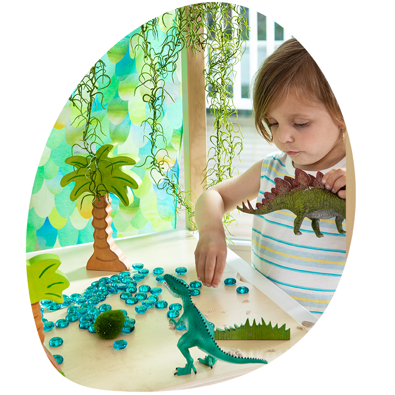 A child plays with toy dinosaurs and blue stones on a table, surrounded by palm tree props and lush green plants.