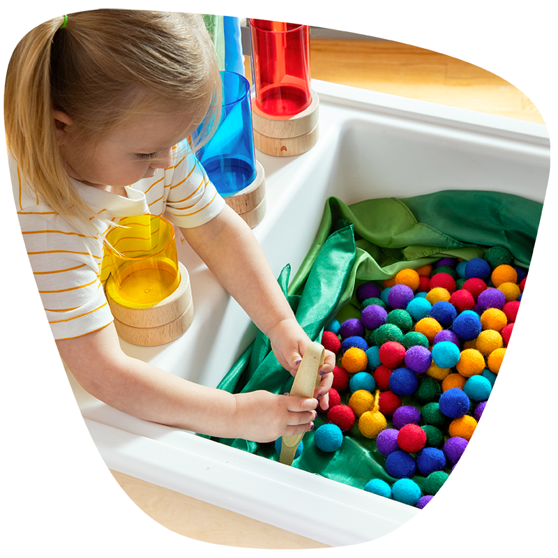 A young girl uses tongs to pick up colorful pom-poms in a sensory bin, sorting them into colored cups nearby for a fun fine motor activity.
