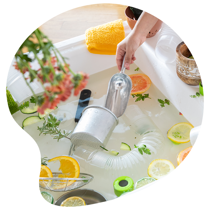 A hand washes dishes in a sink filled with water, citrus slices, herbs, and assorted kitchen items, creating a fresh and aromatic atmosphere.