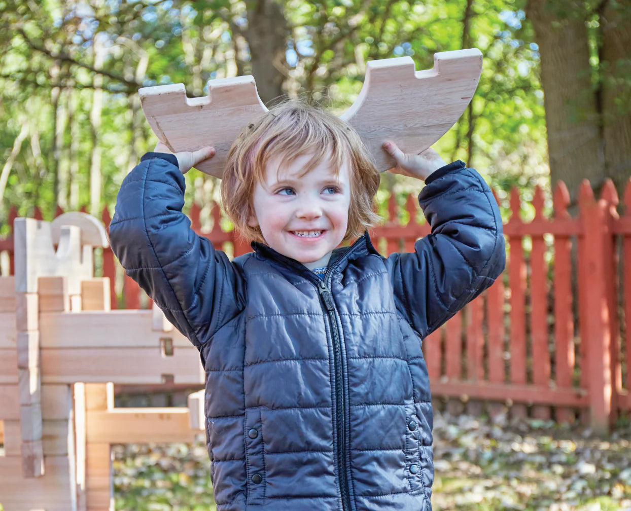 A smiling child in a puffy jacket holds wooden antlers on their head, standing outdoors by a rustic red fence.