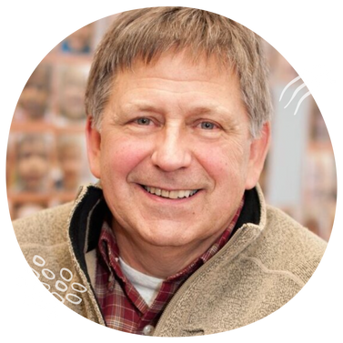 Middle-aged man with short brown hair, dressed in a tan jacket and red plaid shirt, smiles warmly at the camera.