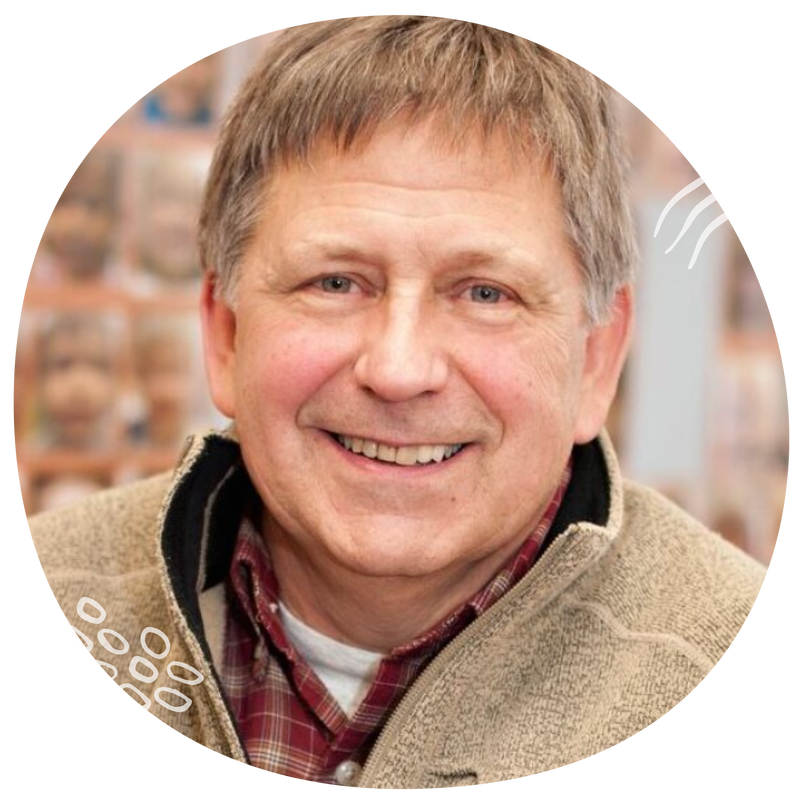 Middle-aged man with short brown hair, dressed in a tan jacket and red plaid shirt, smiles warmly at the camera.
