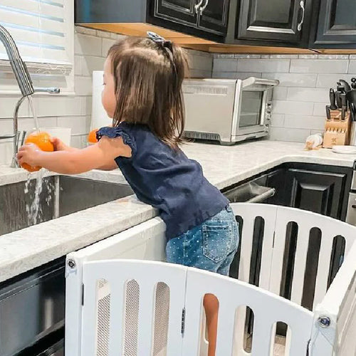 A young girl stands in a playpen at the kitchen sink, carefully washing fresh oranges under running water.