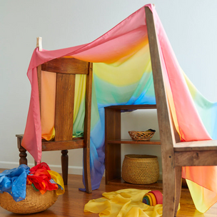 Two wooden chairs and a shelf hold up a Double Playsilk rainbow fort; baskets with open-ended toys and colorful fabric rest on the floor, all made from natural materials.