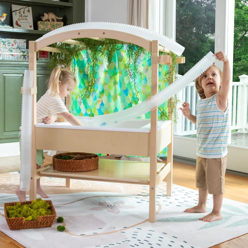 Two young children play with a sensory table and flexible tube in a bright, colorful room filled with natural light from large windows.