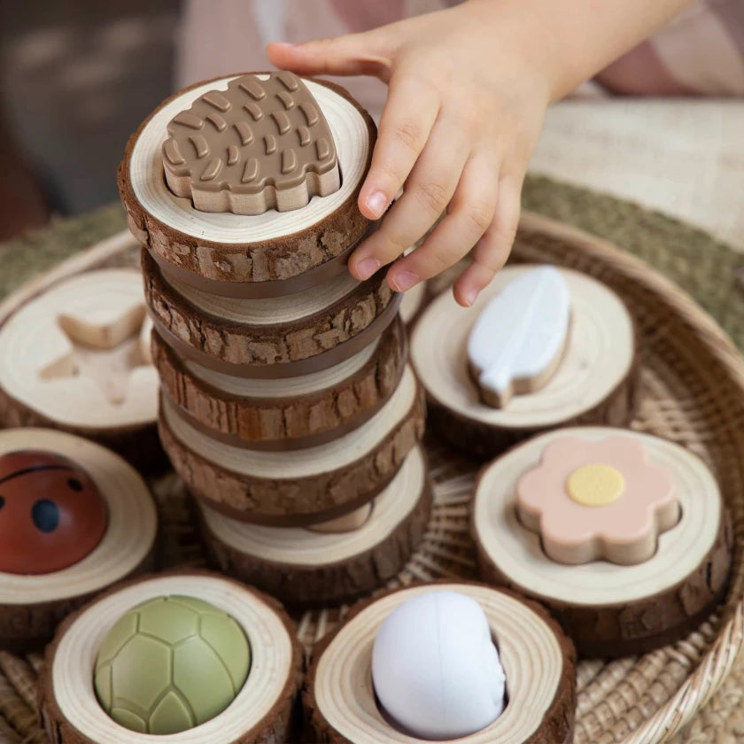 A child’s hand carefully stacks wooden discs featuring nature-themed shapes like a flower, leaf, and turtle shell.