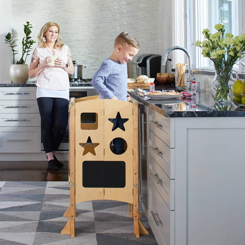 A smiling woman watches a boy stand on a wooden stool at a kitchen counter, surrounded by fresh food and colorful flowers.