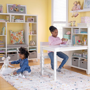 Two young girls play in a bright, organized space filled with shelves, toys, and colorful arts and crafts supplies.